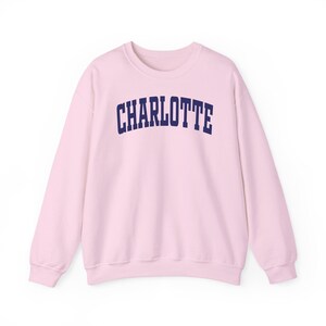 May include: A light pink sweatshirt with the word "CHARLOTTE" printed across the chest in navy blue, arched lettering. The sweatshirt has long sleeves and a crew neck.
