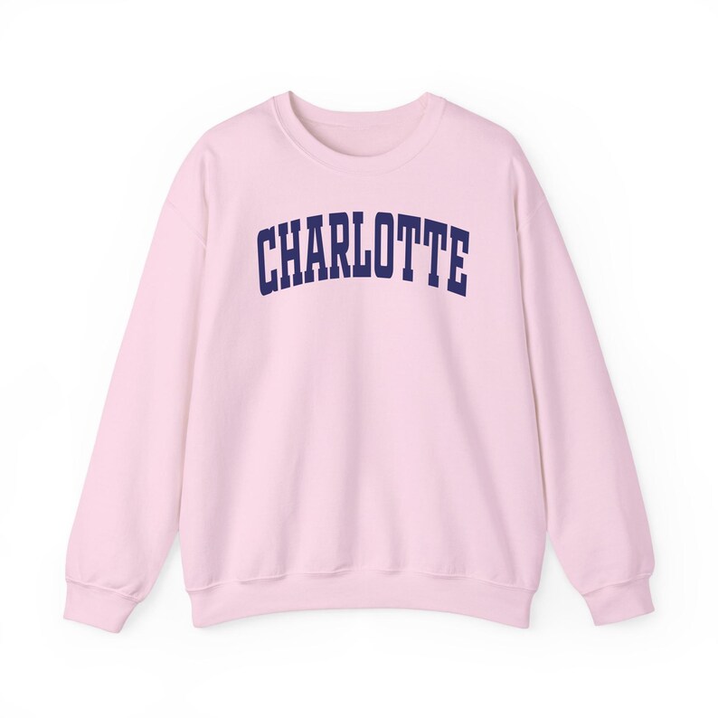 May include: A light pink sweatshirt with the word "CHARLOTTE" printed across the chest in navy blue, arched lettering. The sweatshirt has long sleeves and a crew neck.