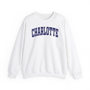 May include: White crew neck sweatshirt with the word "CHARLOTTE" printed in navy blue across the chest. The sweatshirt has long sleeves and a classic, casual design, suitable for everyday wear.
