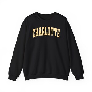 May include: Black crewneck sweatshirt with the word "CHARLOTTE" printed in a gold, collegiate-style font across the chest. The sweatshirt has long sleeves and a classic, casual design, suitable for everyday wear.