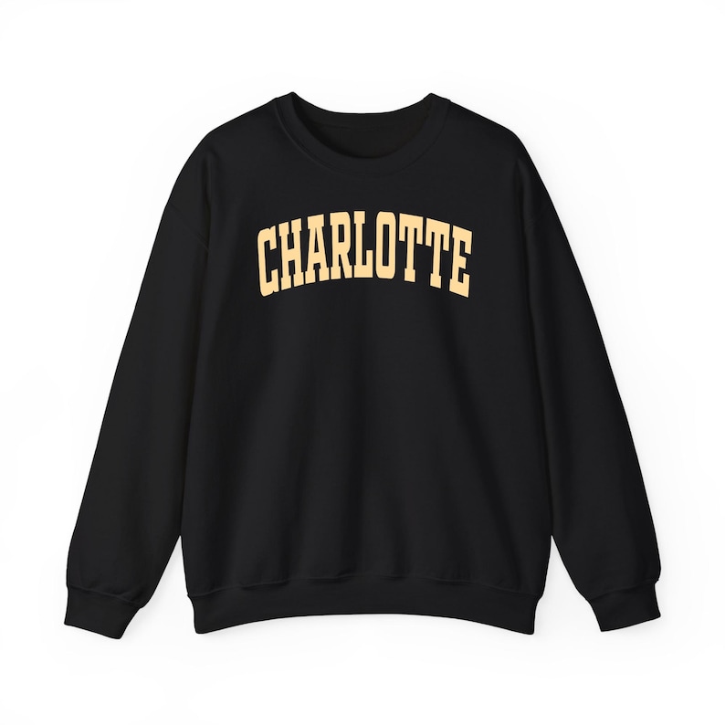 May include: Black crewneck sweatshirt with the word "CHARLOTTE" printed in a gold, collegiate-style font across the chest. The sweatshirt has long sleeves and a classic, casual design, suitable for everyday wear.