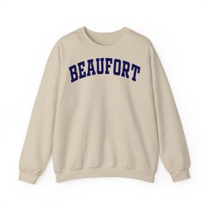 May include: A cream-colored sweatshirt with the word "BEAUFORT" printed across the chest in navy blue, arched lettering. The long-sleeved crewneck top is made of a soft material.