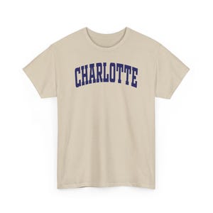 May include: A beige t-shirt with the word "CHARLOTTE" printed across the chest in navy blue, arched lettering. The shirt has short sleeves and a crew neck.
