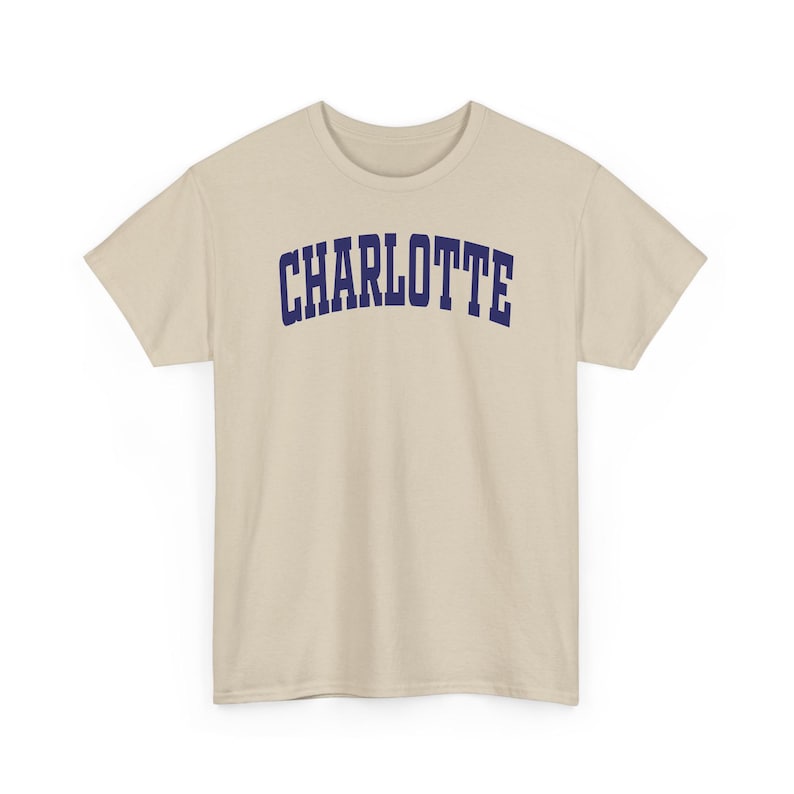 May include: A beige t-shirt with the word "CHARLOTTE" printed across the chest in navy blue, arched lettering. The shirt has short sleeves and a crew neck.
