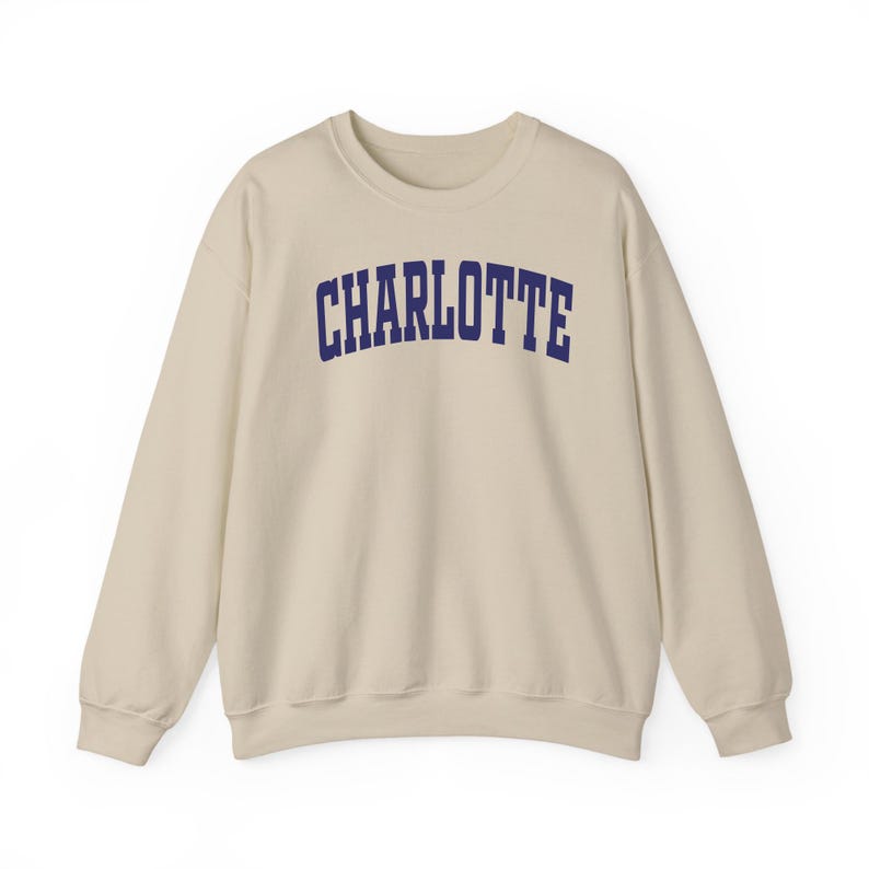 May include: A cream-colored sweatshirt with the word "CHARLOTTE" printed across the chest in navy blue, block letters. The long-sleeved top is made of a soft material and has a crew neck.