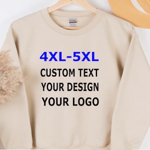 May include: A beige sweatshirt with the text "4XL-5XL CUSTOM TEXT YOUR DESIGN YOUR LOGO" in blue and black. The sweatshirt is hanging on a wooden hanger.