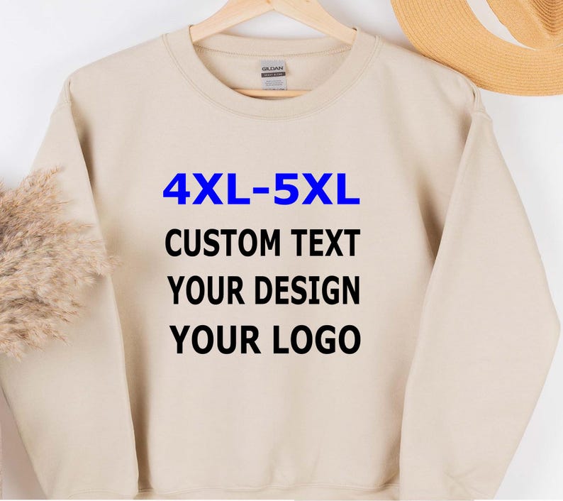 May include: A beige sweatshirt with the text "4XL-5XL CUSTOM TEXT YOUR DESIGN YOUR LOGO" in blue and black. The sweatshirt is hanging on a wooden hanger.