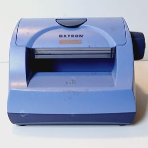 May include: A blue and navy Xyron 1500 machine. The machine has a curved top with a black handle and the brand name and model number are printed on the front. The machine has a slot for inserting materials.