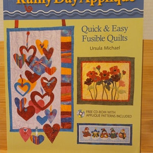 May include: Book cover for "Rainy Day Appliqué: Quick & Easy Fusible Quilts" by Ursula Michael. The cover features a quilt with hearts and a quilt with flowers. The book includes a free CD-ROM with appliqué patterns.