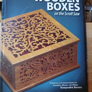 May include: A book cover titled "Creating Wooden Boxes on the Scroll Saw" features a detailed wooden box with a decorative, openwork design. The box is light brown with a contrasting dark red interior. The cover also includes text about patterns and instructions for jewelry, music, and keepsake boxes.