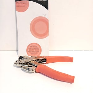 May include: A We R Memory Keepers Crop-A-Dile tool with coral-colored handles and a silver metal body. The tool is displayed in front of its original white and black packaging, which states it punches 1/8 inch and 3/16 inch holes and sets eyelets.