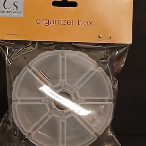 May include: A clear, round organizer box with multiple compartments, packaged in a clear plastic bag. The packaging features the text "organizer box" and the Crafter's Square logo.