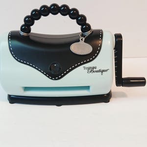 May include: A light blue and black Texture Boutique die-cutting machine with a black beaded handle and a silver charm. The machine has a black crank and a black base. The text "Texture Boutique" is printed on the front.