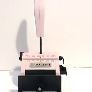 May include: A light pink Zutter Innovative Products binding machine featuring a vertical handle, a black base with a central adjustment knob, and a series of rectangular slots along the top edge for aligning paper or covers during the binding process.