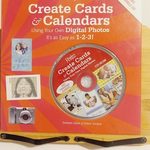 May include: A red and orange book titled "Create Cards & Calendars" with a CD-ROM featuring a photo of a child. The book cover features the Reader's Digest logo and the text "Using Your Own Digital Photos. It's as Easy as 1-2-3!" The CD-ROM is labeled "Create Cards & Calendars" and features the Reader's Digest logo. The CD-ROM is labeled "Using Your Own Digital Photos" and "CD-ROM".