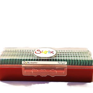 May include: A red plastic storage box with a clear lid, filled with green craft supplies. The box has a clear plastic clasp and a white label with the word "Sizzix" in a colorful logo. The box is labeled "Sizzix Sizzlits" and "Pop Squad".