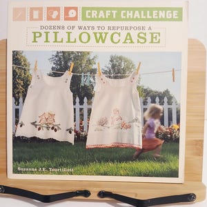 May include: A book titled "Craft Challenge" with the subtitle "Dozens of Ways to Repurpose a Pillowcase." The cover shows two white dresses with embroidery hanging on a clothesline. A child runs in the background. The author's name is Suzanne J.E. Tourtillott.