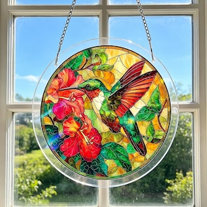 May include: A round stained glass window hanging with a hummingbird and hibiscus flowers. The hummingbird is green, red, and orange, with a long beak. The flowers are red and pink, with green leaves. The window is hanging from a chain.