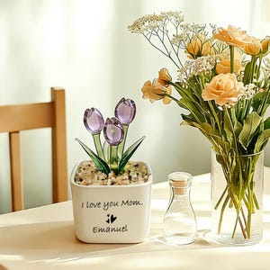 May include: A white ceramic pot with the text "I love you Mom." and "Emanuel" in black script. The pot contains three purple glass tulip flowers. A vase of orange flowers and a small glass bottle are also present.
