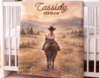 May include: A personalised baby blanket with a landscape illustration of a child riding a horse. The blanket displays the name "Cassidy" and the word "Grace" in a script font. The background depicts a sunset over rolling hills.