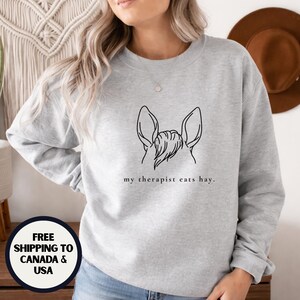 Op de afbeelding: Lichtgrijze sweatshirt met een zwarte lijntekening van de oren en manen van een paard. De tekst "my therapist eats hay." staat eronder. De sweatshirt heeft een ronde hals.