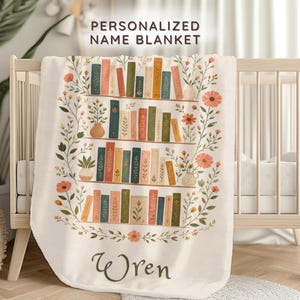 May include: Cream-colored personalized baby blanket with a bookshelf design. The blanket features a floral border and the name "Wren" in script. The bookshelf design includes colorful books, vases, and flowers. The text "PERSONALIZED NAME BLANKET" is at the top.