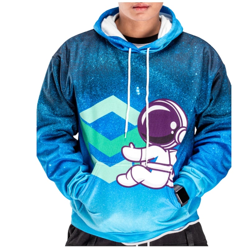 Pode incluir: Um moletom com capuz azul com uma estampa degrad&ecirc; inspirada em uma gal&aacute;xia, um capuz com cord&atilde;o branco e um gr&aacute;fico de um astronauta de desenho animado sentado ao lado de uma grande forma geom&eacute;trica em chevron verde-claro.