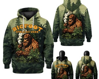 May include: A green hooded sweatshirt featuring an all-over forest print with a graphic of Bigfoot smoking a pipe amidst cannabis plants. The text "BIGFOOT STOLE MY WEED" is printed in orange above the creature. The design appears on both sides.