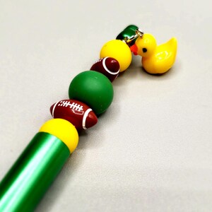 Puede incluir: Un bolígrafo verde con una cuenta amarilla y verde, una cuenta de fútbol marrón y un amuleto de pato de goma amarillo.