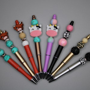 May include: Eight colorful pens with different animal and patterned beads. The pens have gold, silver, and black barrels. The beads are turquoise, brown, white, pink, black, and leopard print.