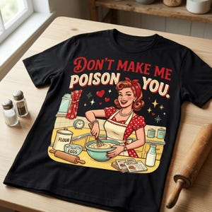 以下が含まれることがあります： 「Don't Make Me Poison You」のテキストが入った、女性が料理をしているレトロなイラストが描かれた黒いTシャツ。デザインには、ベーキング材料と麺棒のあるキッチンシーンが含まれています。女性は赤い水玉模様のドレスとエプロンを着ています。