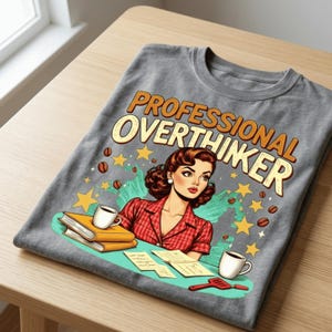 以下が含まれることがあります： オレンジとクリーム色の文字で「PROFESSIONAL OVERTHINKER」と書かれたグレーのTシャツ。グラフィックは、赤いチェックのシャツを着た女性のヴィンテージスタイルのイラストで、コーヒーカップ、本、星に囲まれています。