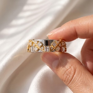 May include: A wide band ring featuring a polished silver-tone base overlaid with a decorative gold-tone metal lattice. The gold design is set with multiple clear, square-cut princess stones and smaller round accent stones arranged in a pattern.