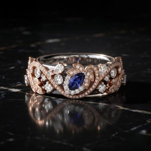 May include: A rose gold ring featuring a central marquise-cut blue sapphire surrounded by small round white diamonds. The band is intricately detailed with a vine-like pattern of rose gold leaves and additional round white diamonds throughout.