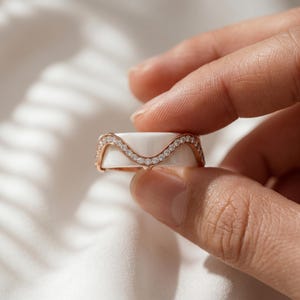 Peut inclure: Une bague en or rose composée d'un anneau en céramique blanche lisse avec une incrustation centrale ondulée. Les bords en or rose sont sertis d'une rangée continue de petites pierres rondes, claires et scintillantes qui suivent la courbe.