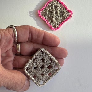 May include: Two handmade crochet earrings. One earring has a gray center with a bright pink border. The other earring is gray with a cream-colored border. Both earrings are square-shaped and have a silver loop for hanging.