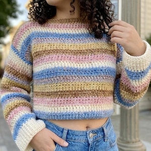 May include: A cropped, crochet sweater with horizontal stripes in pastel colors: light blue, pink, yellow, and brown. The sleeves and neckline are a cream color. The sweater is paired with blue denim jeans.