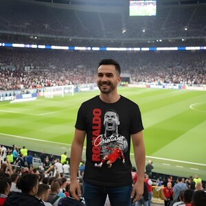 May include: A black t-shirt featuring a graphic of Cristiano Ronaldo in action, with the name "Ronaldo" in red vertical text and "Cristiano" in white script. The person is standing in a stadium, wearing blue jeans.