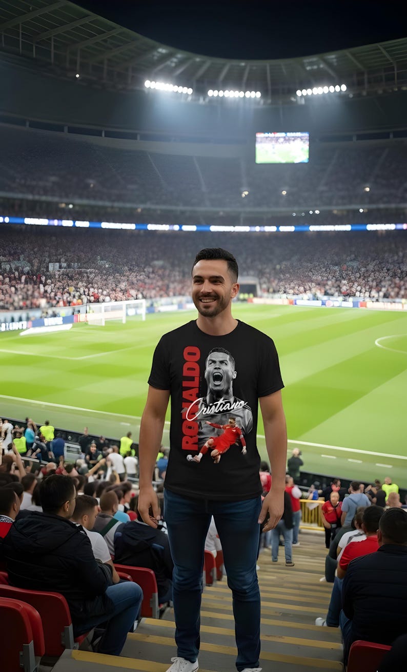 May include: A black t-shirt featuring a graphic of Cristiano Ronaldo in action, with the name "Ronaldo" in red vertical text and "Cristiano" in white script. The person is standing in a stadium, wearing blue jeans.