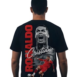 May include: Black t-shirt featuring a large image of Cristiano Ronaldo. The design includes a portrait of Ronaldo with the name "Cristiano" in cursive and the word "RONALDO" in red vertical text. A second image shows Ronaldo in a red uniform.