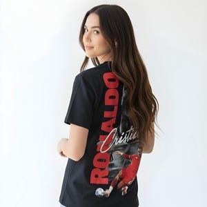 May include: Black t-shirt featuring a graphic of a soccer player in a red uniform, with the name "Ronaldo" in red vertical text. The shirt is worn by a person with long brown hair and light blue jeans.