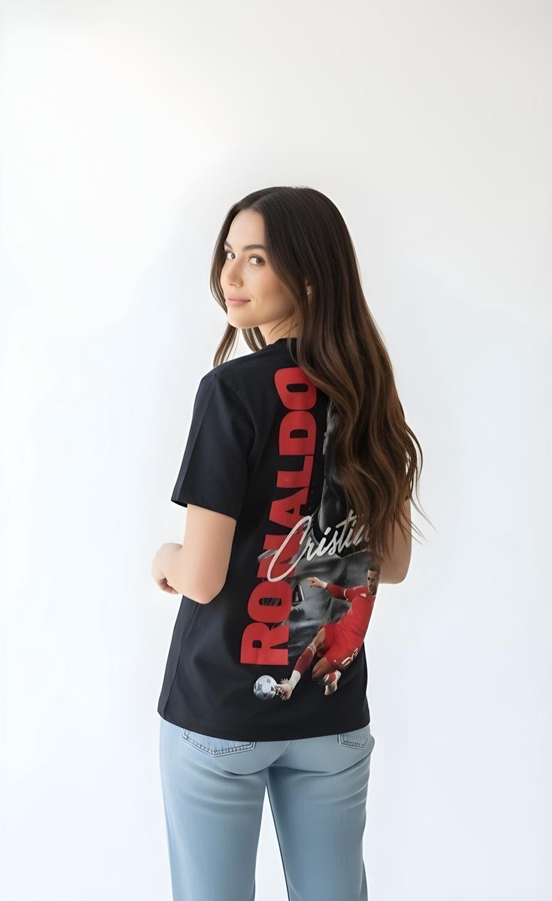 May include: Black t-shirt featuring a graphic of a soccer player in a red uniform, with the name "Ronaldo" in red vertical text. The shirt is worn by a person with long brown hair and light blue jeans.