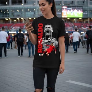May include: Black t-shirt featuring a graphic of Cristiano Ronaldo with the name "Ronaldo" in red vertical text. The shirt also displays the name "Cristiano" in a cursive font. The person is wearing black jeans and white sneakers.