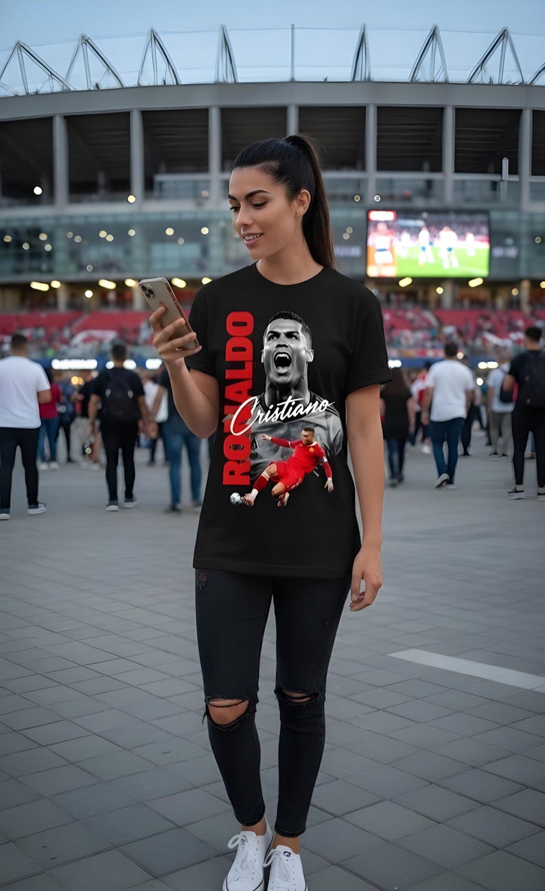 May include: Black t-shirt featuring a graphic of Cristiano Ronaldo with the name "Ronaldo" in red vertical text. The shirt also displays the name "Cristiano" in a cursive font. The person is wearing black jeans and white sneakers.