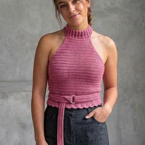 Puede incluir: Top halter de ganchillo rosa con cuello alto y cinturón. El top tiene un borde festoneado y un cinturón a juego con hebilla plateada. La modelo lleva pantalones grises oscuros.