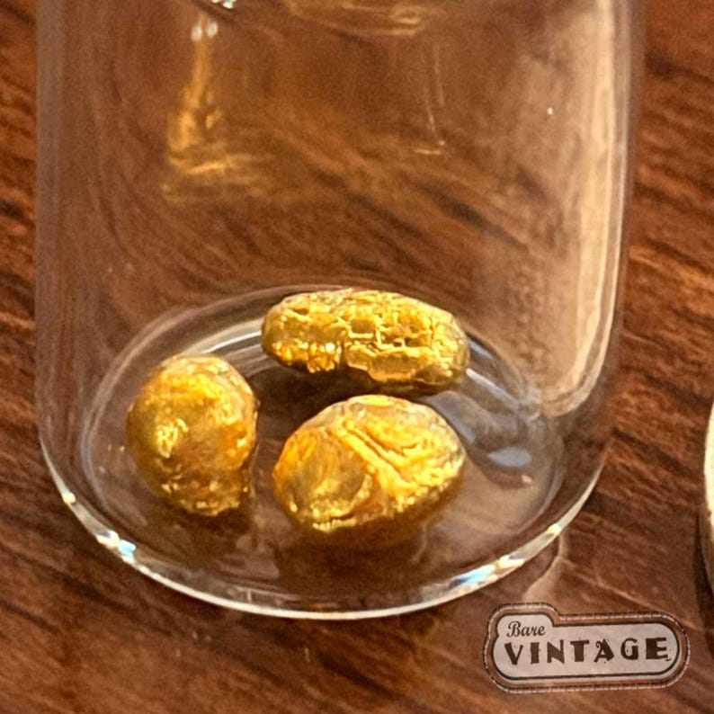Op de afbeelding: Een heldere glazen pot met drie goudkleurige nuggets. De nuggets hebben een textuur. De pot heeft een kleine, heldere basis. Het label onderaan luidt "Bare Vintage".