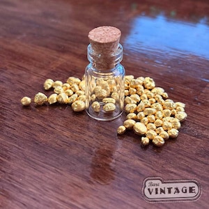 Op de afbeelding: Een kleine, heldere glazen fles met een kurk, gevuld met kleine, goudkleurige nuggets. De fles staat op een houten oppervlak, omringd door meer van de gouden nuggets. De afbeelding is goed verlicht, waardoor de textuur en glans van het goud worden benadrukt.