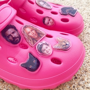 May include: A pair of bright pink foam clogs featuring multiple custom photo charms attached to the upper surface. The charms include portraits of various people, a black cat, and a spotted feline, all secured within the ventilation holes of the shoes.