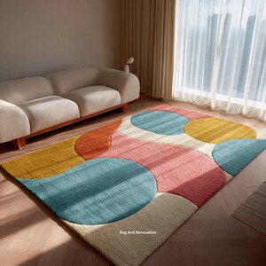 May include: A colorful area rug with a modern design. The rug features large, overlapping shapes in shades of blue, yellow, orange, pink, and cream. The text "Rug And Renovation" is visible in the bottom right corner.