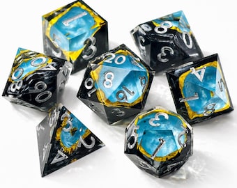May include: A set of seven polyhedral dice featuring a translucent blue core encased in black resin with gold leaf accents. Each die has sharp edges and engraved silver numbers, including a d20, d12, d10, d8, d6, d4, and a percentile die.